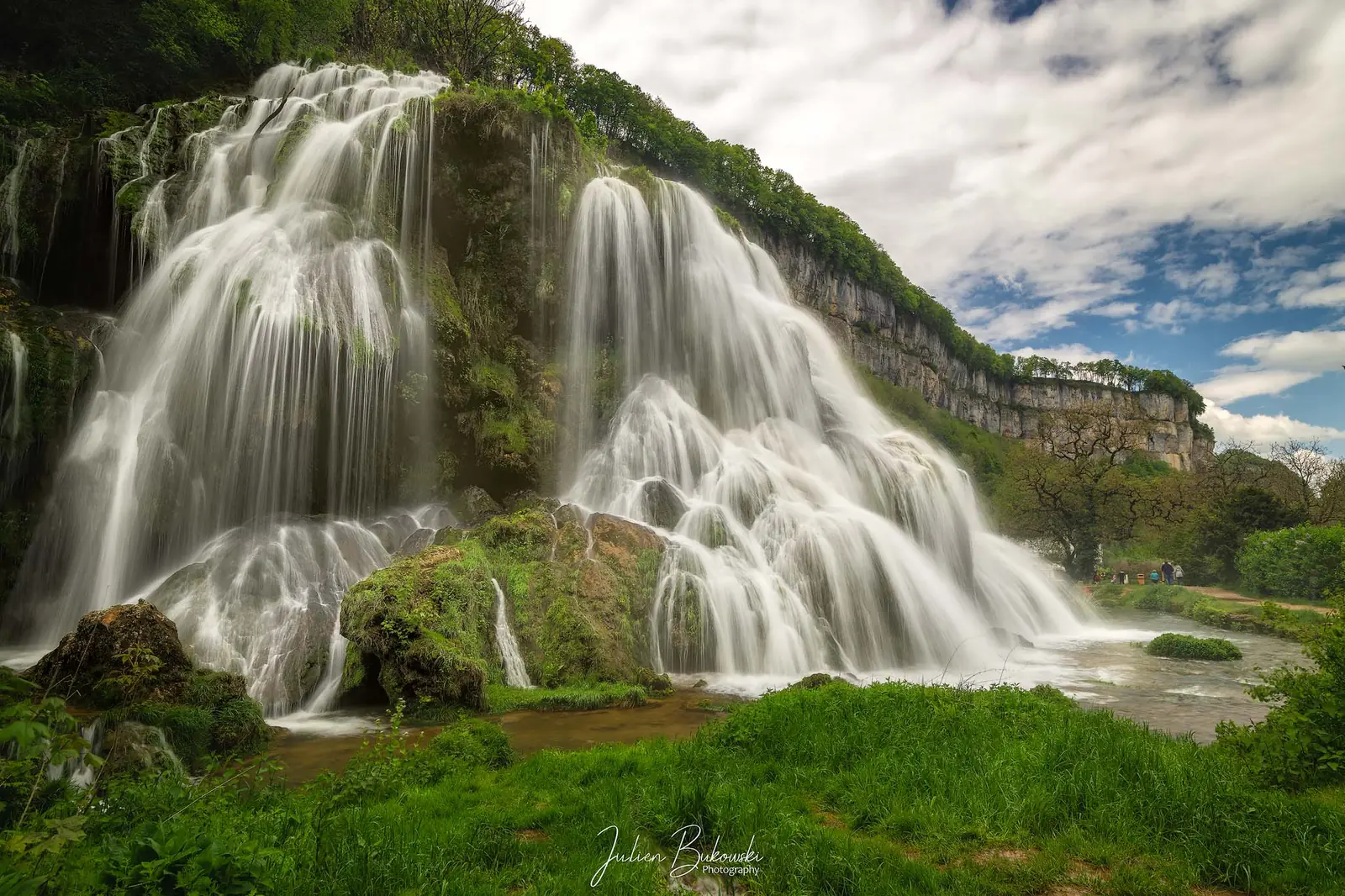 Large cascade en voile qui ruisselle sur des tufs moussus, avec une pelouse verte au premier plan. Falaises et ciel de nuages au fond.