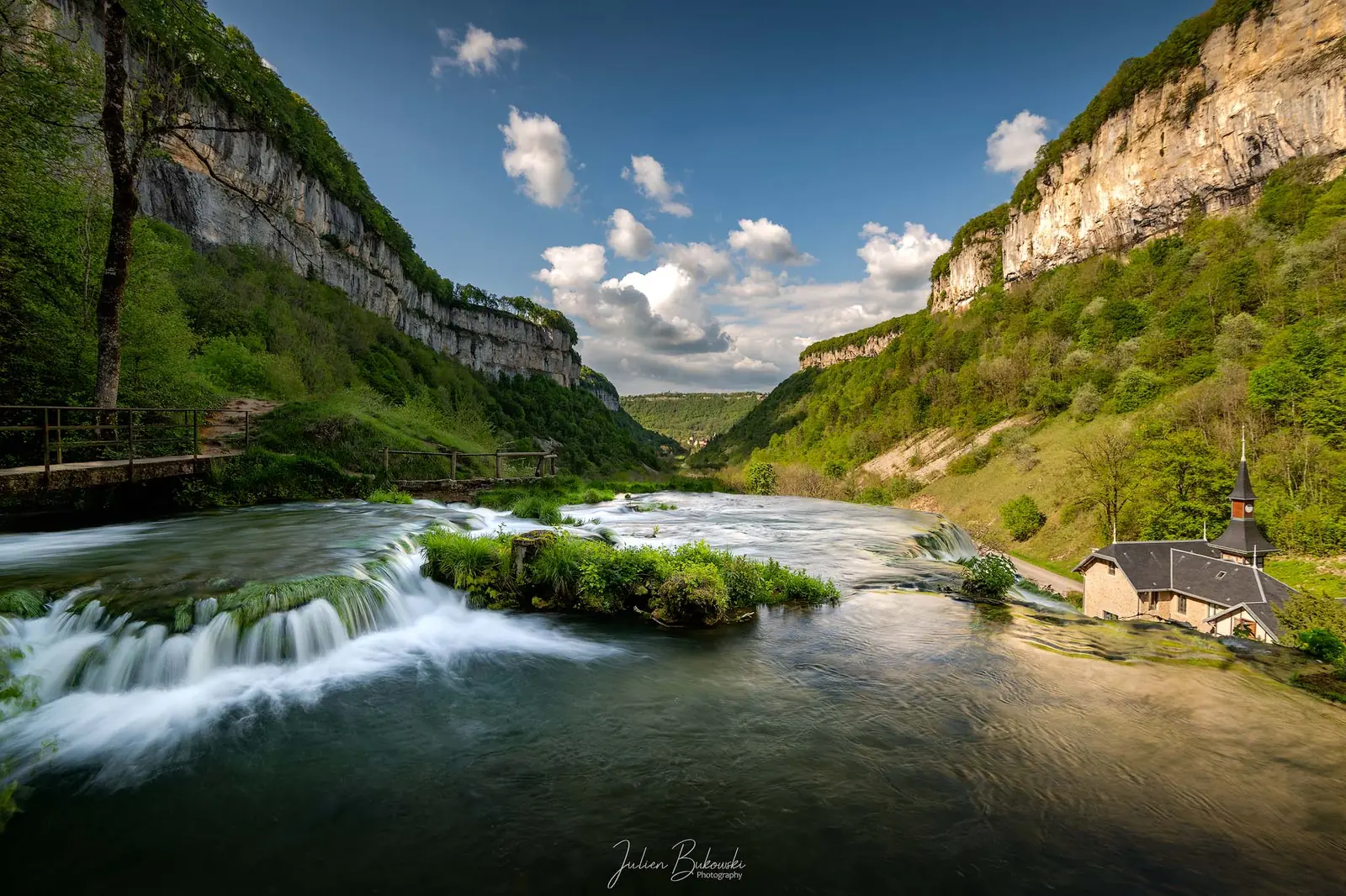 Rivière en petits seuils au fond d'une vallée encaissée entre deux falaises calcaires. Une chapelle à clocher se trouve à droite, sous un ciel bleu à nuages de printemps.