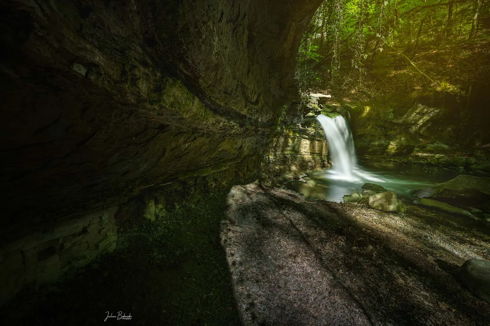 Vue prise depuis un abri sous roche sombre : une dalle inclinée au premier plan, la chute qui brille au fond dans la lumière.