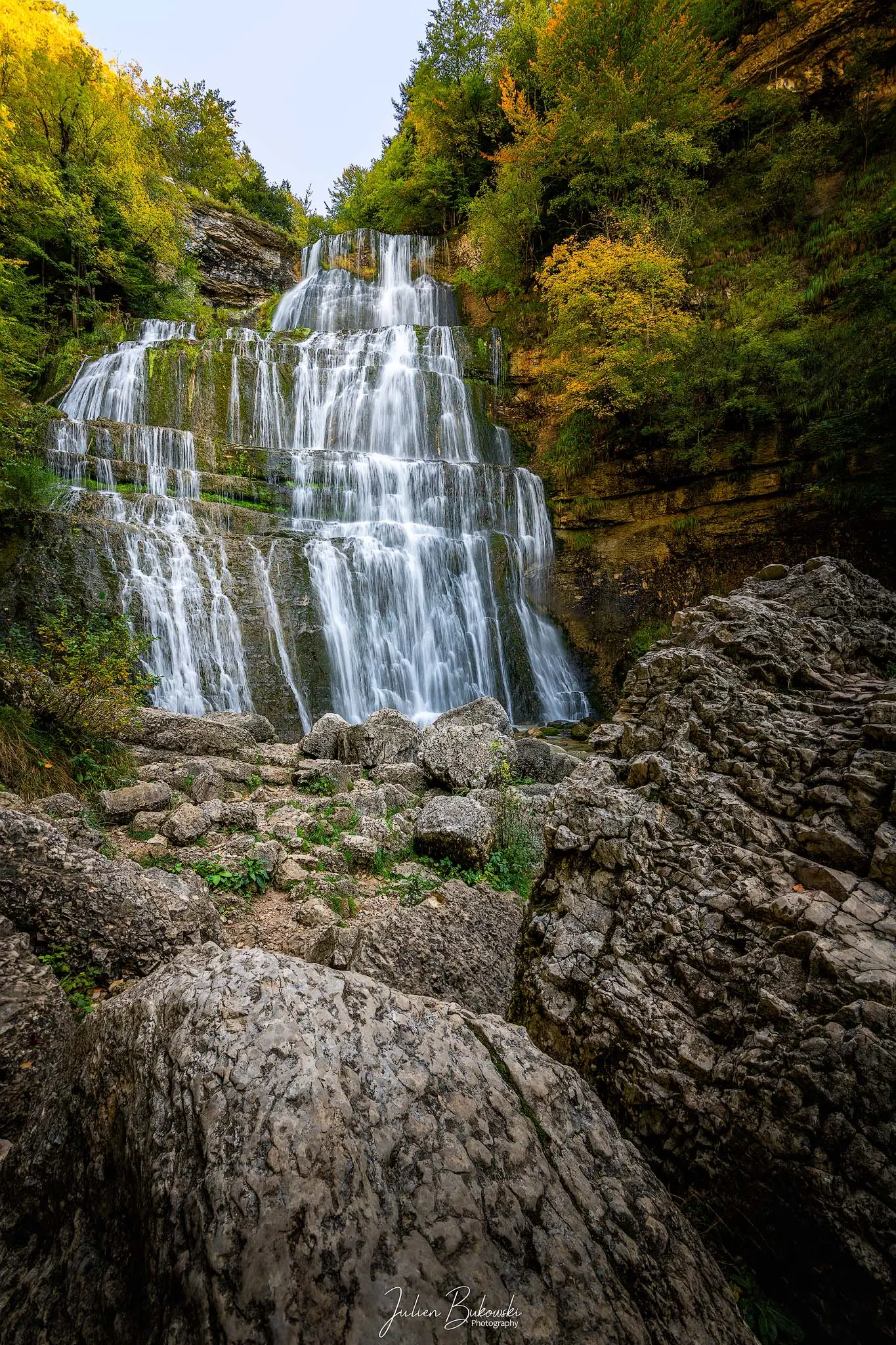 Haute cascade en gradins encadrée de feuillus qui jaunissent, avec de gros blocs calcaires au premier plan.