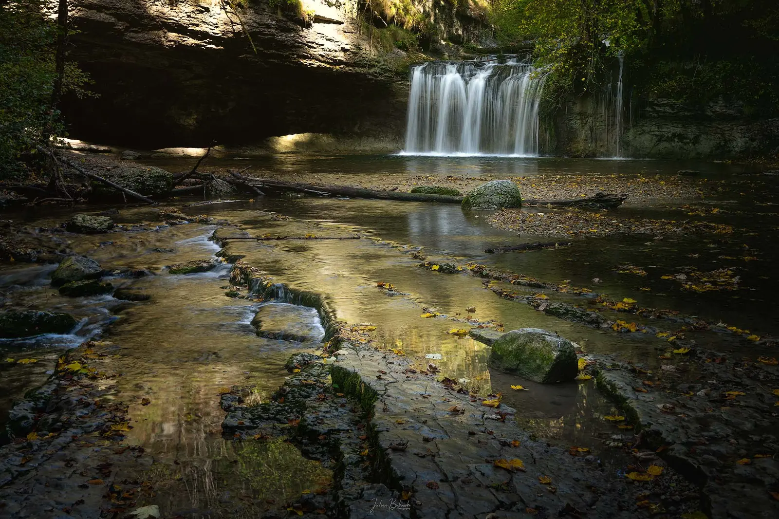 Chute en rideau au fond d'un cirque de roche sombre. Au premier plan, des dalles mouillées et des feuilles éparses brillent dans une lumière basse.