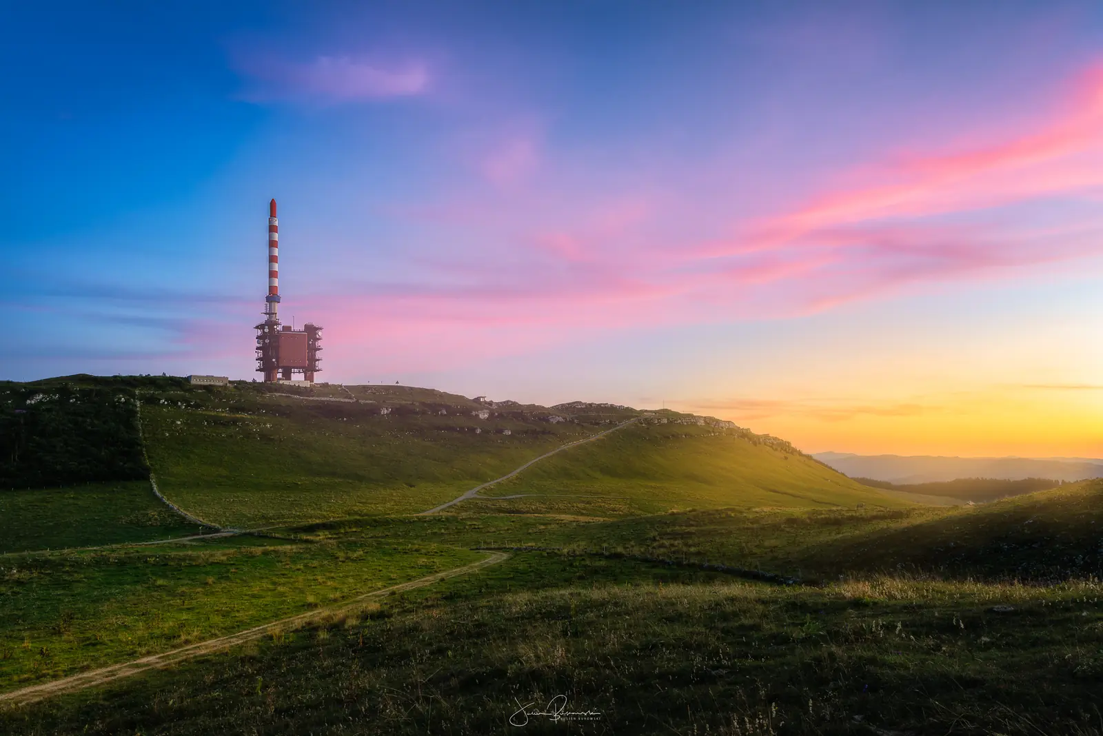 Crête herbeuse traversée de sentiers, surmontée d'une haute antenne, sous un ciel rose et orange de coucher de soleil d'été.