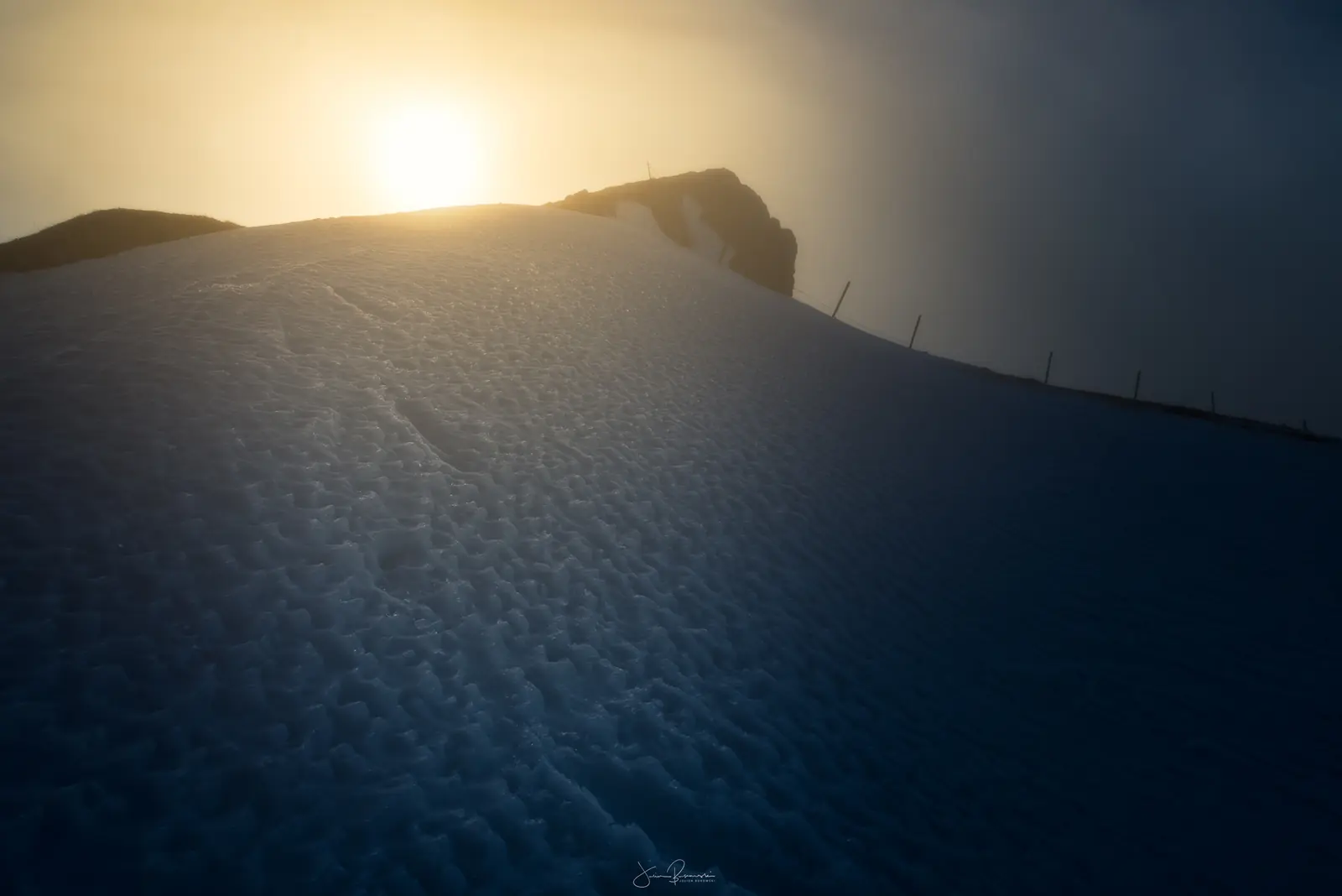 Pente de neige bleutée et marquée de traces, avec une ligne de piquets. Le soleil bas perce une brume dorée juste au-dessus de la crête.