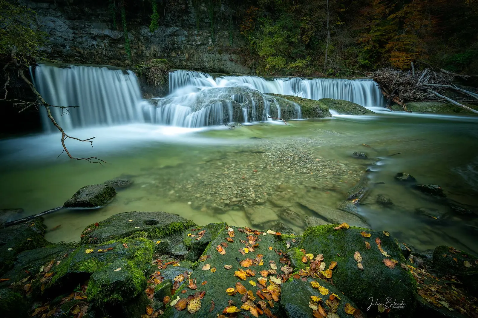 Chute en rideau qui se jette dans une vasque verte et claire. Le premier plan est fait de rochers moussus couverts de feuilles orange tombées. Automne.