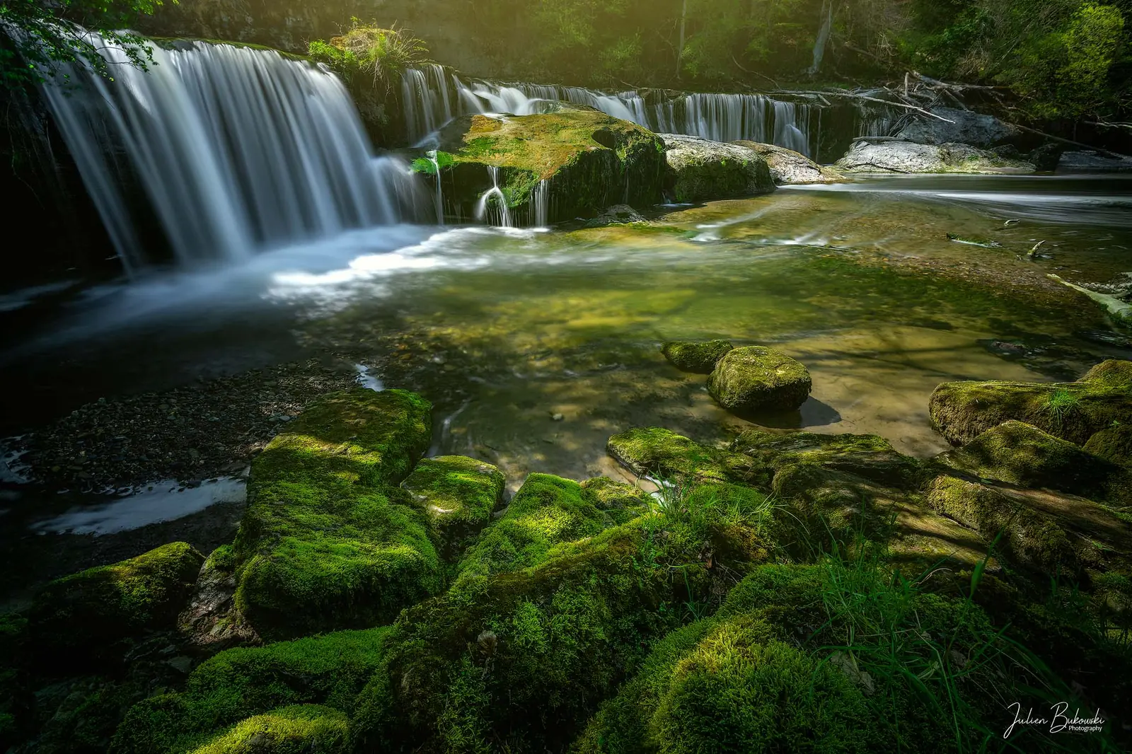 Chute basse sur une dalle, avec une succession de blocs couverts de mousse vert vif au premier plan. Lumière d'après-midi de printemps.
