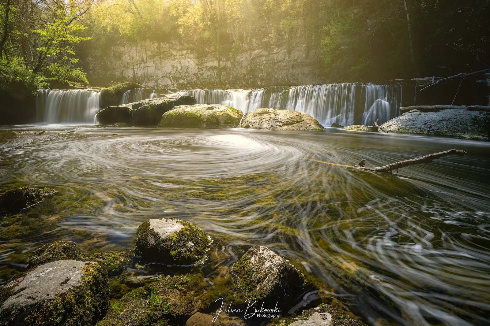 Large chute basse en rideau. Au premier plan, l'écume dessine des tourbillons en pose longue autour de blocs. Lumière dorée et brumeuse qui passe à travers les arbres.