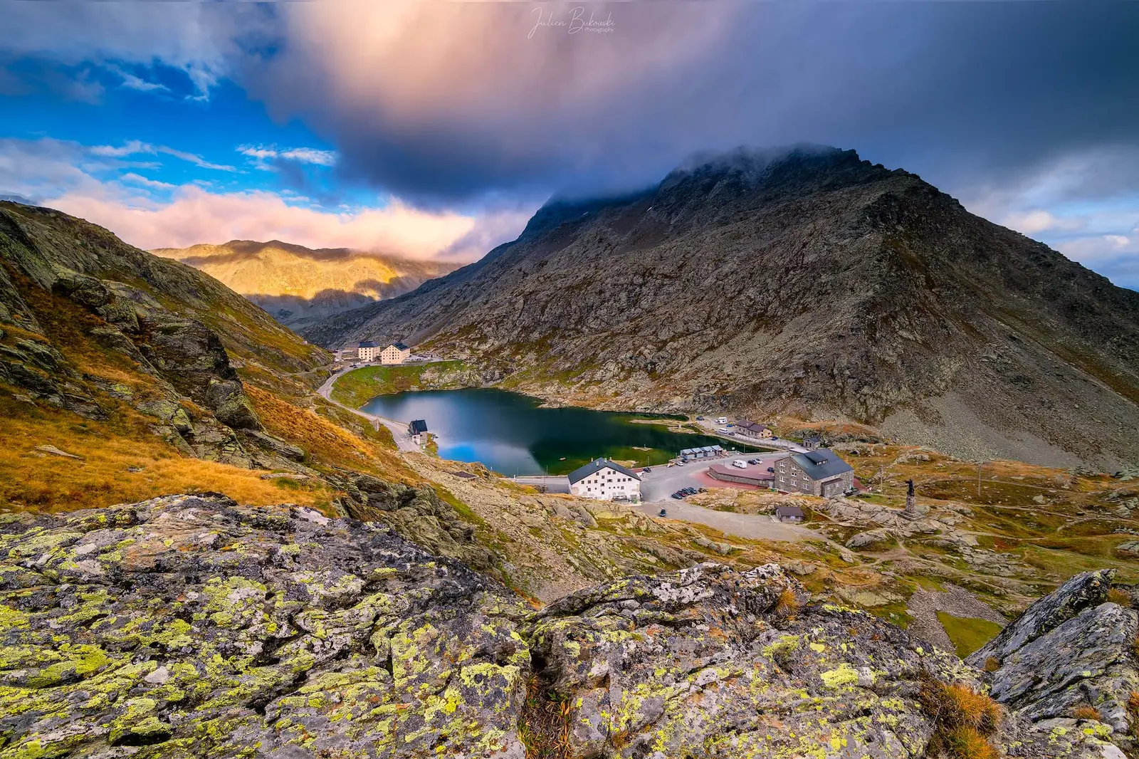 Vue plongeante sur un petit lac de col bordé de bâtiments. Rochers couverts de lichen et herbes rousses au premier plan, nuages bas sur le sommet. Fin d'été.