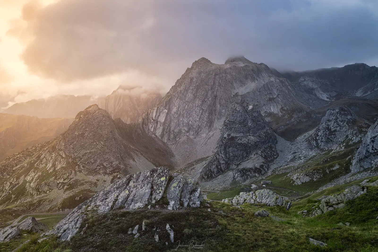Sommets rocheux sous des nuages qui s'accrochent, avec un contre-jour doré à gauche. Pâturages et dalles claires au premier plan.