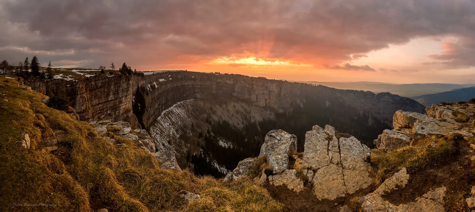 Tout l'arc du cirque au coucher du soleil, sous un ciel lourd : le soleil perce les nuages au-dessus de la paroi du fond, quelques plaques de neige sur la crête.