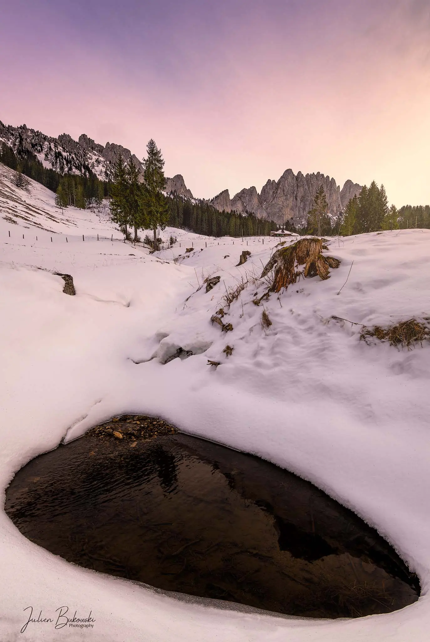 Trou d'eau sombre dans la neige au premier plan. Au fond, une crête dentelée et quelques sapins sous un ciel rose de fin de journée.