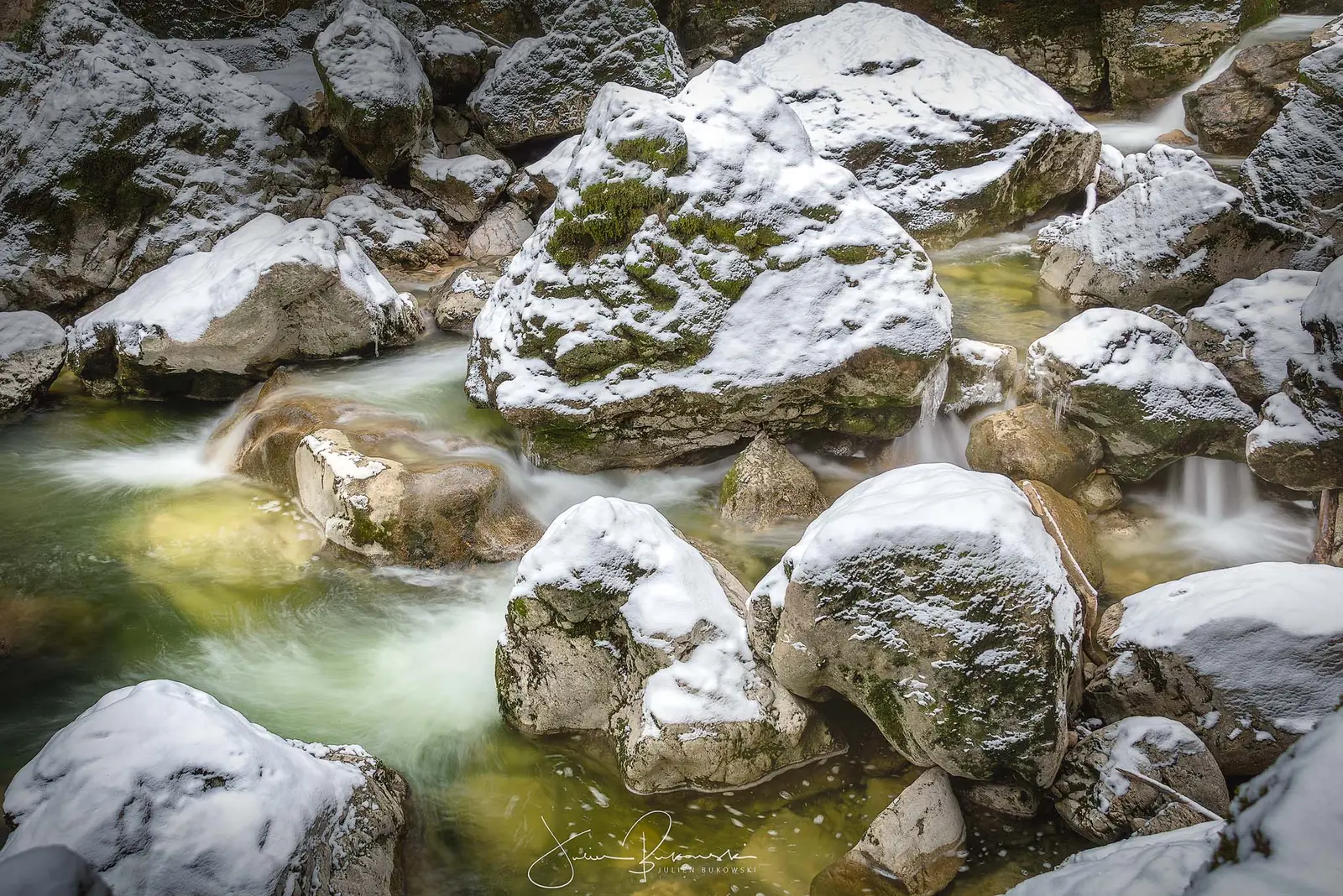 Gros blocs de rocher coiffés de neige au milieu d'une eau verte, rendue soyeuse par la pose longue. Scène d'hiver.