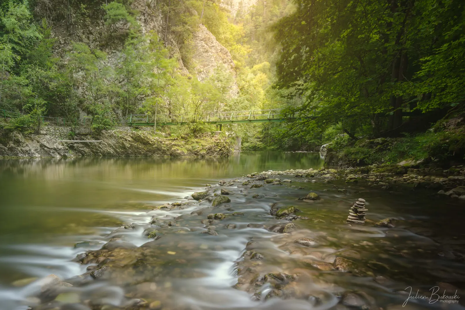 Rivière large et calme, en pose longue, qui file sur un seuil de galets. Une passerelle traverse au fond, sous un soleil de fin de journée qui éclaire les feuillus d'été.
