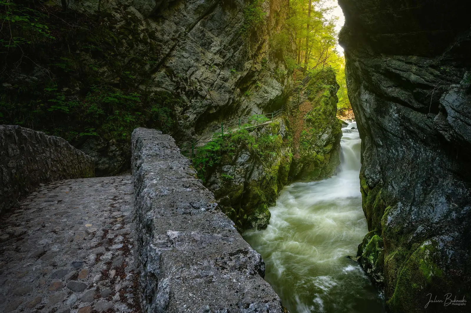 Chemin pavé bordé d'un parapet de pierre, qui longe un torrent blanc serré entre deux parois sombres. La végétation est verte au printemps, avec une lumière douce de fin d'après-midi.