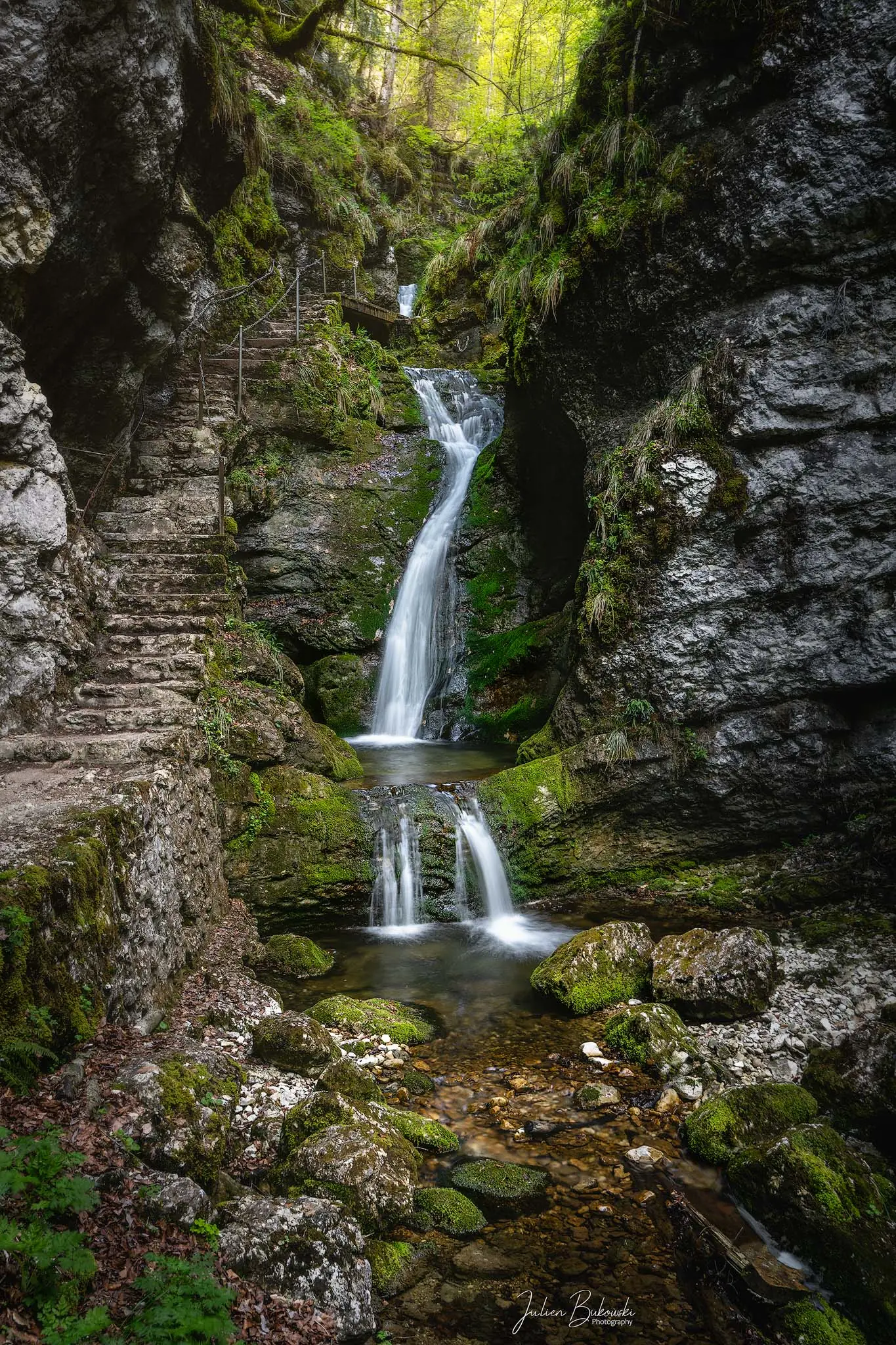Cascade à deux étages dans une gorge étroite. Un escalier de pierre est taillé dans la paroi à gauche, et la mousse couvre les blocs au premier plan. Feuillage vert tendre de printemps.