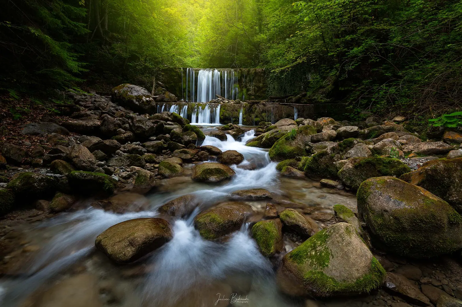 Lit de torrent jonché de gros blocs moussus. L'eau descend en filets vers le spectateur et une chute à gradins se dresse au fond, sous des arbres. Fin d'été.