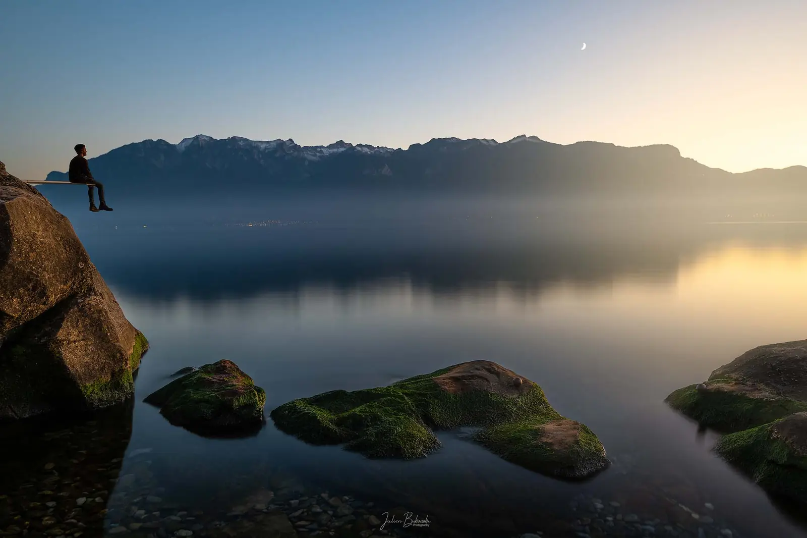 Silhouette assise au bord d'un gros rocher, au-dessus du lac lisse. Les montagnes se découpent en face dans un ciel de crépuscule, avec un fin croissant de lune.