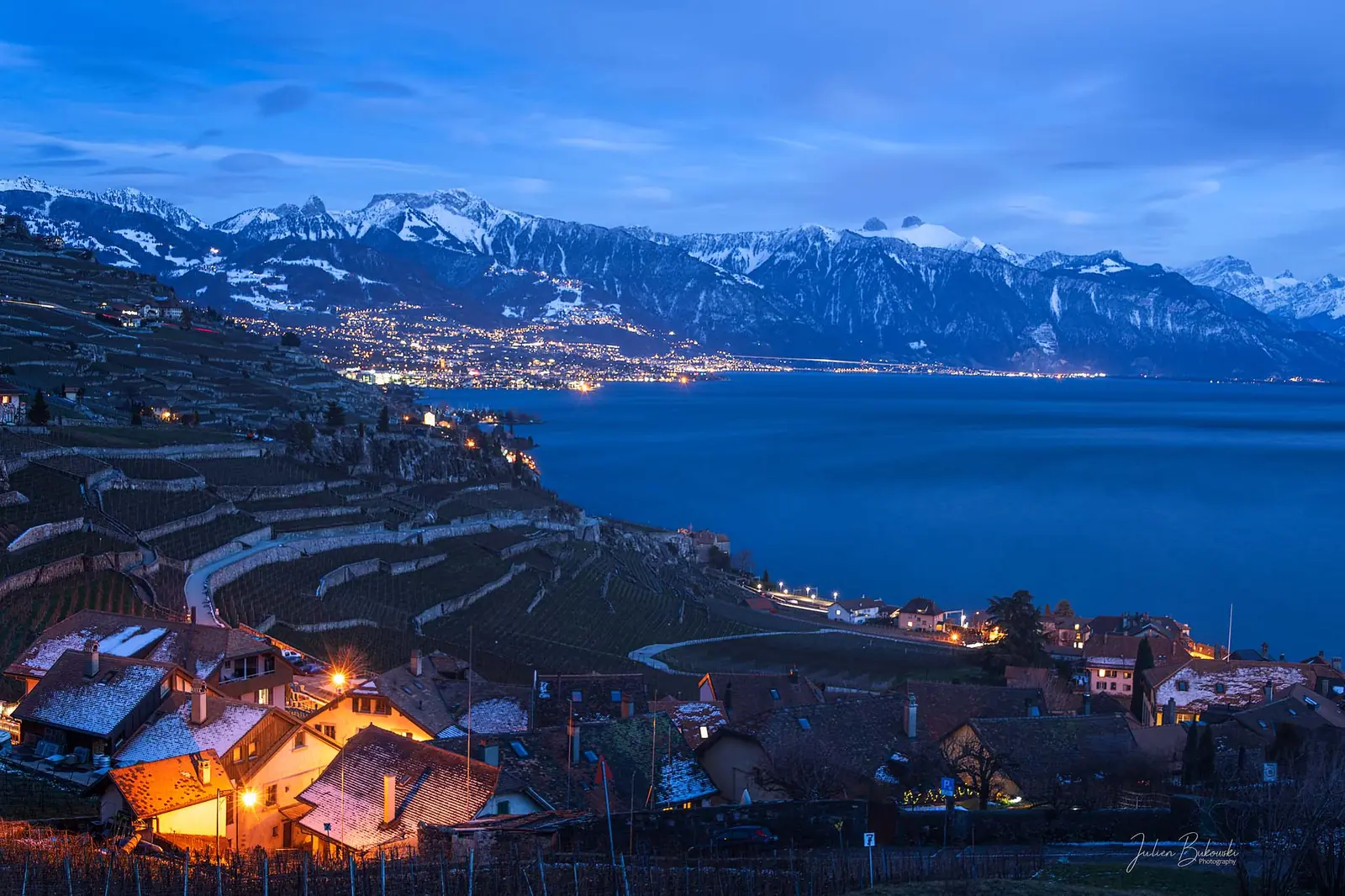 À l'heure bleue, en hiver, les toits éclairés d'un village vigneron au premier plan. Les terrasses de vignes descendent vers le lac, avec une rive illuminée et des montagnes enneigées en face.