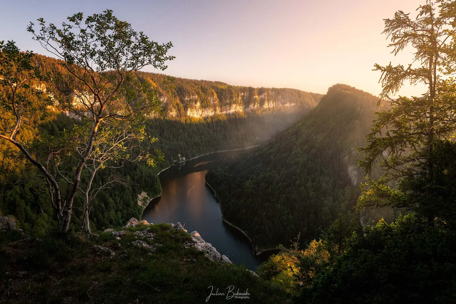 Vue depuis un éperon boisé sur une rivière encaissée entre de hautes falaises, au coucher du soleil. Arbres en silhouette au premier plan.