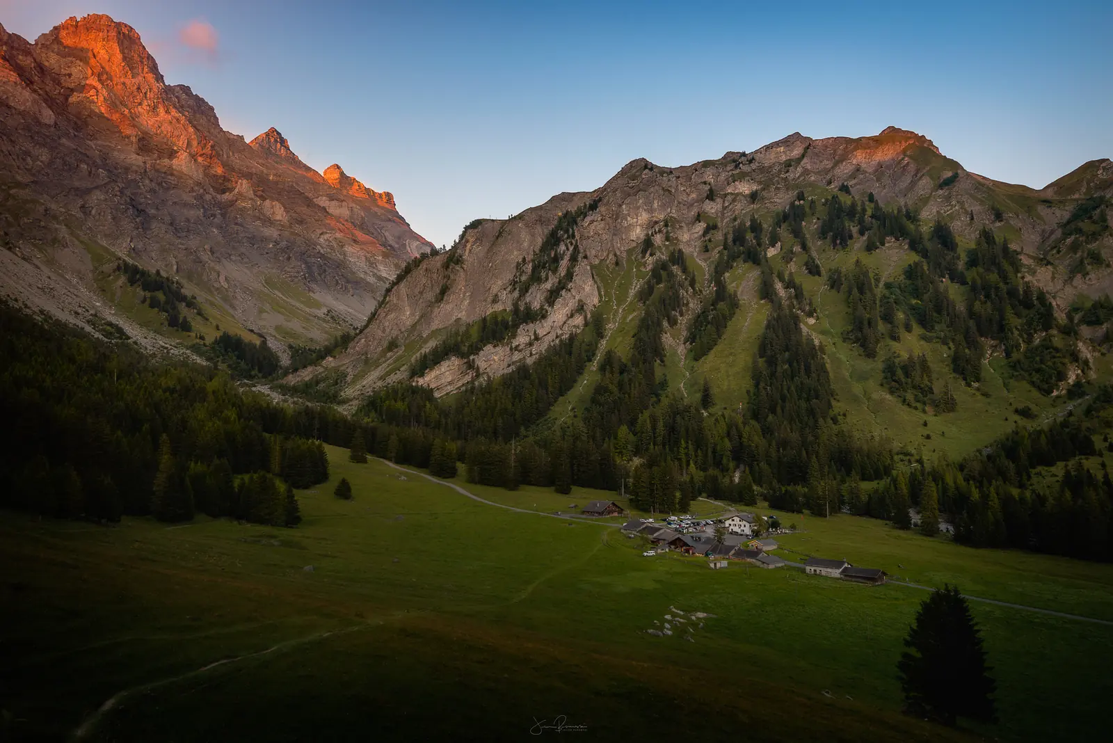 Alpage vert avec un petit hameau de chalets au fond du vallon. Le dernier soleil rougit le sommet rocheux à gauche, et des sapins sont au premier plan.